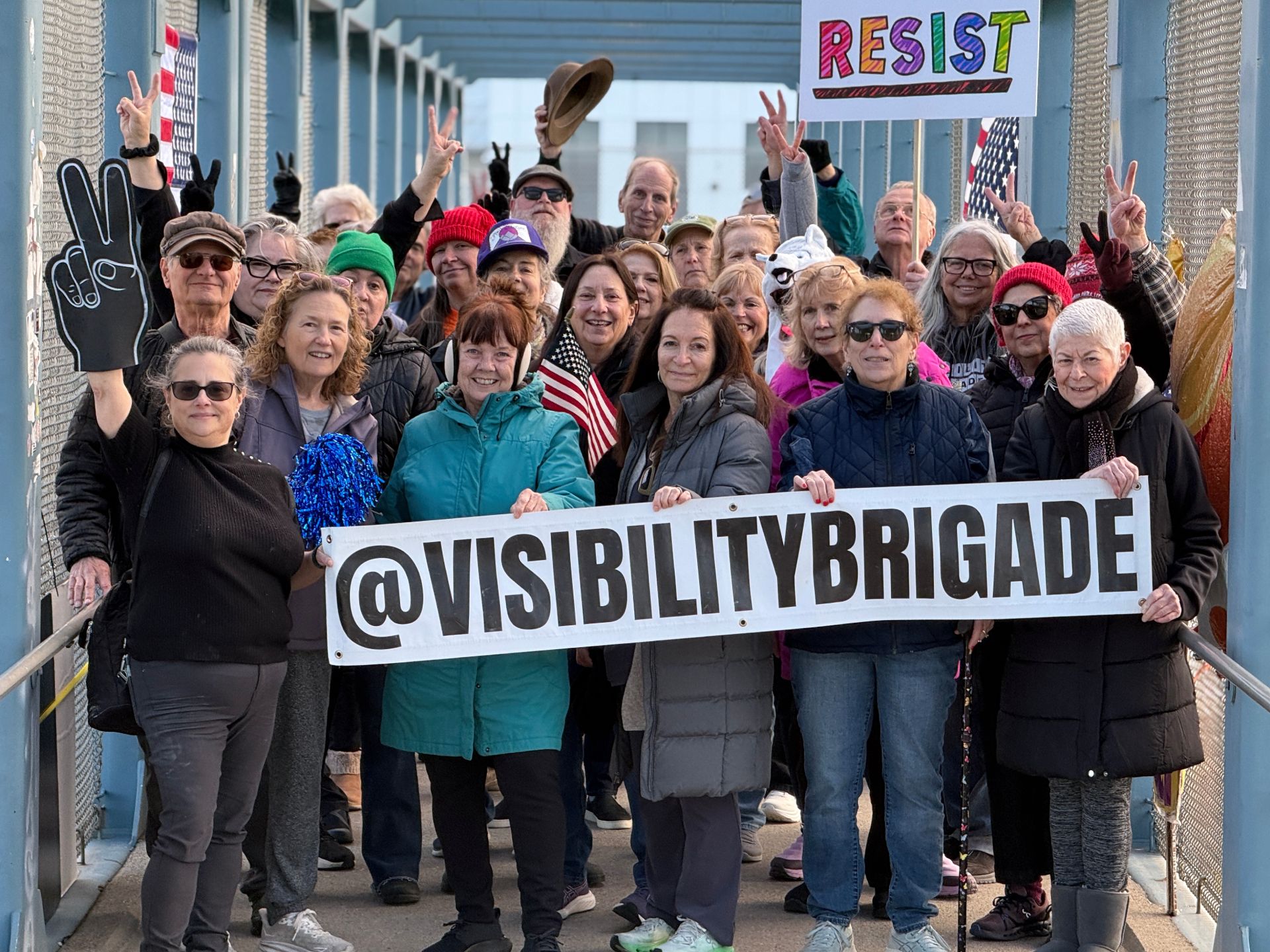 A group of happy protesters standing on a highway overpass smiling and making peace signs with both their hands as well as a foanm finger