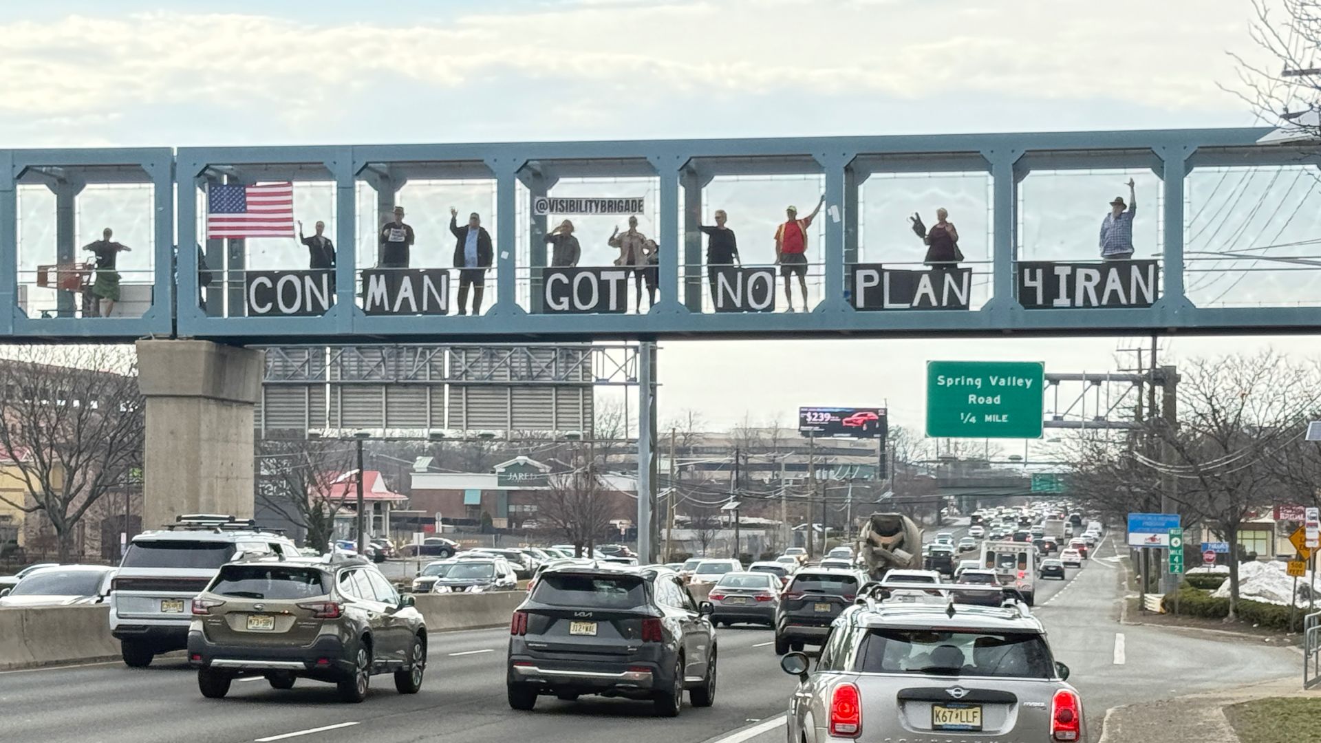 Protesters standing on a highway overpass holding a sign that says 'Con Man Got No Plan'