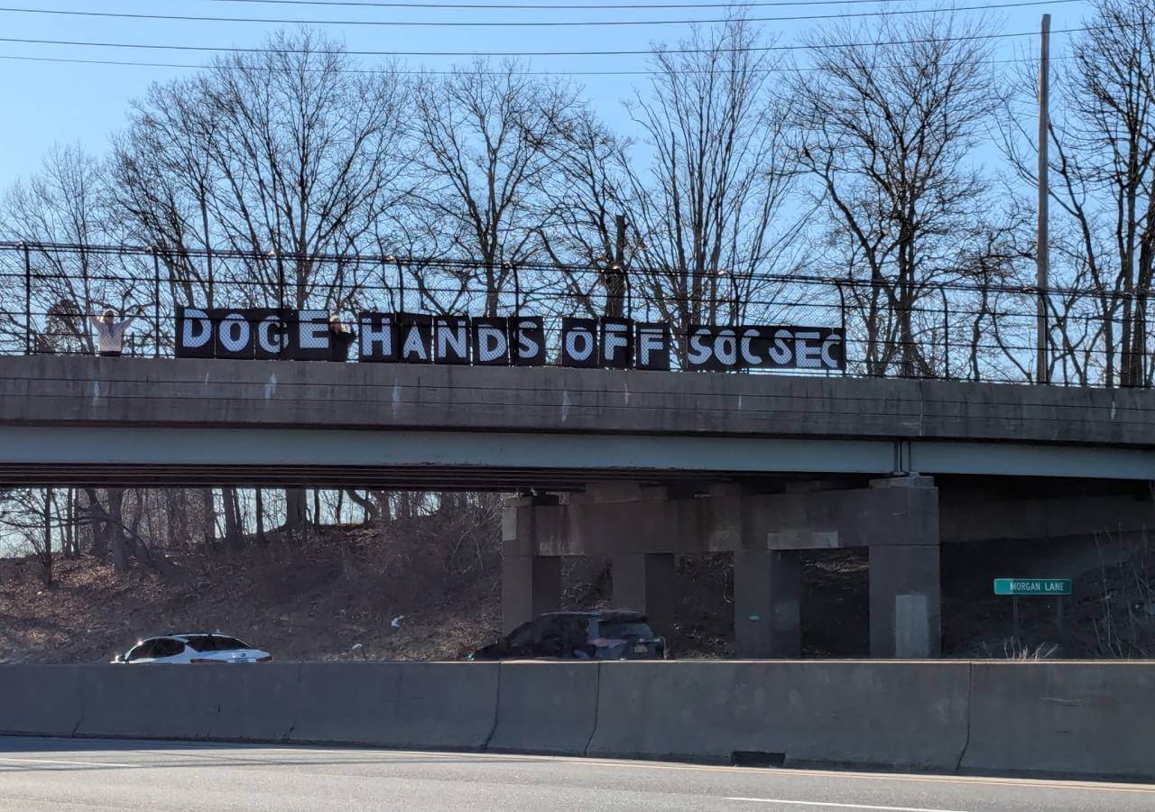 Protesters standing on a highway overpass holding a sign that says 'DOGE Hands Off Soc Sec'