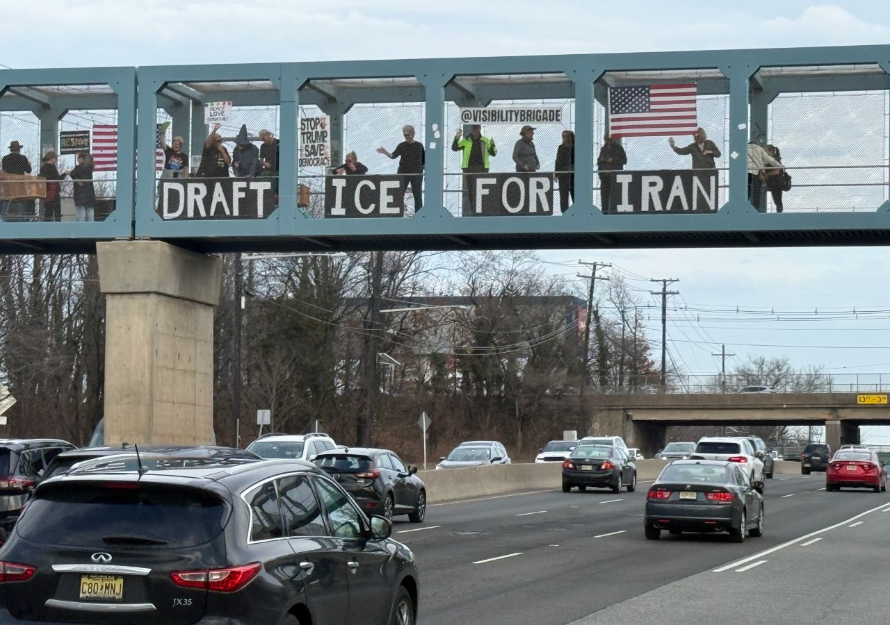 Protesters standing on a highway overpass holding a sign that says 'Draft ICE For Iran'
