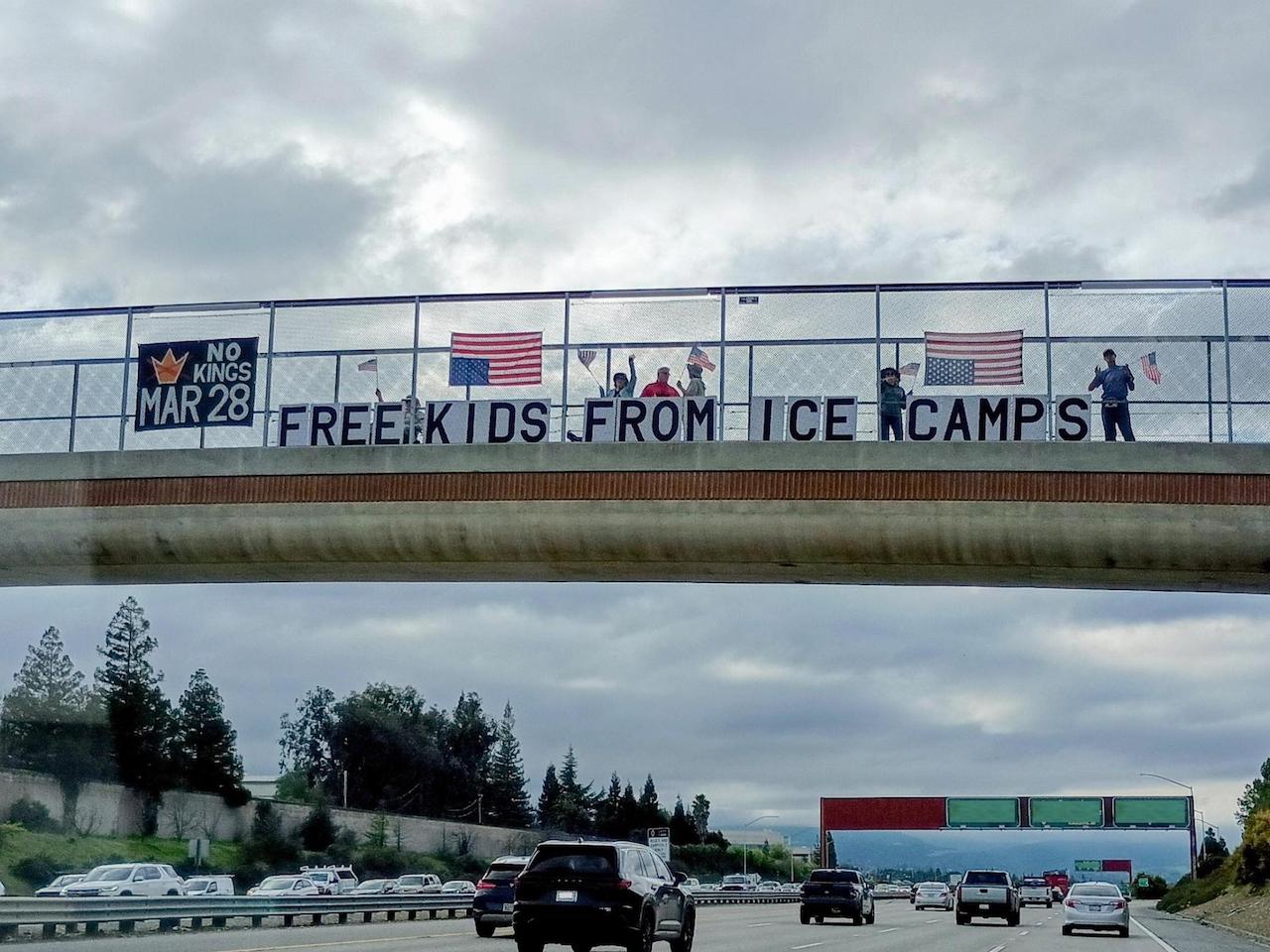 Protesters standing on a highway overpass holding a sign that says 'Free Kids from ICE Camps'