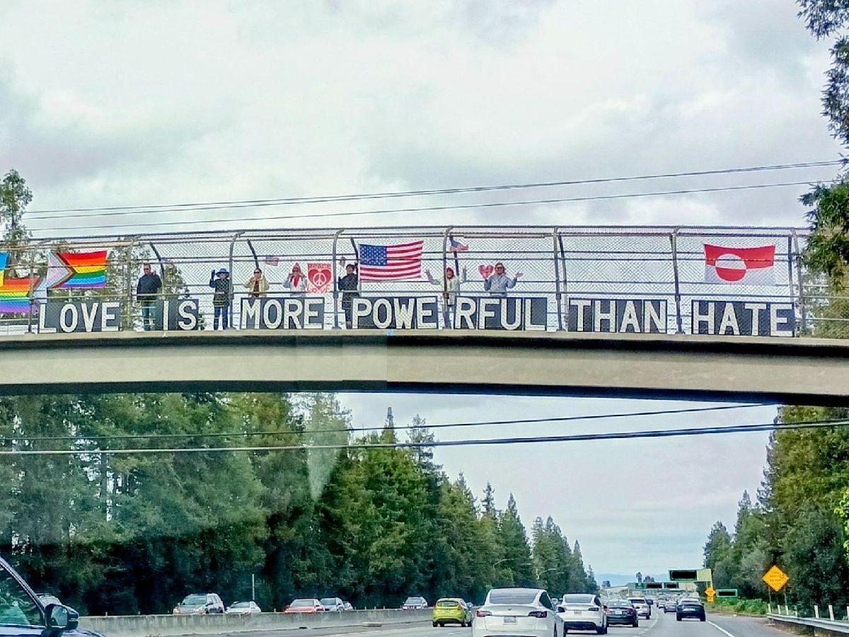 Protesters standing on a highway overpass holding a sign that says 'Love Is More Powerful Than Hate'