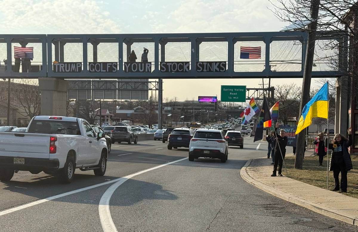 Protesters standing on a highway overpass holding a sign that says 'Trump Golfs Your Stock Sinks'