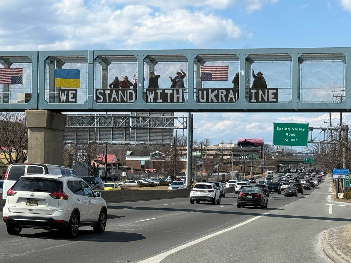 Protesters standing on a highway overpass holding a sign that says 'We Stand With Ukraine'